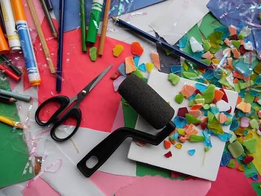 Colorful assortment of craft supplies scattered across a table, including crayons, pencils, scissors, colored paper, a foam paint roller, and small pieces of multicolored craft material.