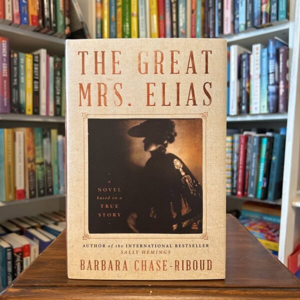 A copy of The Great Mrs. Elias by Barbara Chase-Riboud stands upright on a wooden table in front of softly blurred library bookshelves. The beige book cover features the title in large brown lettering above a sepia-toned photograph of a woman in profile wearing a wide-brimmed hat. Text on the cover describes it as “a novel based on a true story,” and notes that the author also wrote the international bestseller Sally Hemings. The library setting in the background emphasizes the book as a featured reading recommendation.
