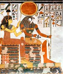 Ancient Egyptian wall painting depicting two seated figures in profile. A woman on the left raises one hand toward a larger figure seated on the right, who has the head of a bird and wears traditional Egyptian garments and jewelry. Hieroglyphic symbols fill the background, and a large red-orange sun disk appears above the figures. The artwork features the flat, stylized forms and earth-tone colors typical of ancient Egyptian tomb paintings.
