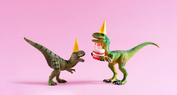 Two toy dinosaurs wearing yellow party hats face each other against a pink background. The larger green dinosaur holds a small white birthday cake with red frosting and lit candles, while the smaller brown dinosaur appears to look up at the cake. The playful scene creates a festive birthday celebration with dinosaur toys.