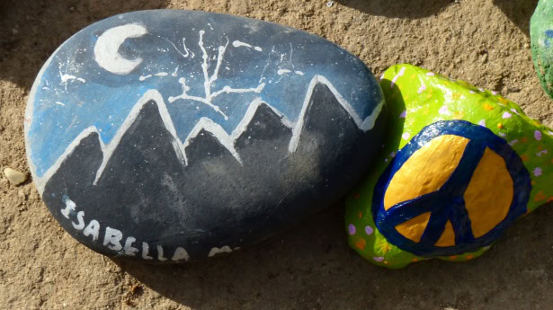 Two painted rocks rest on sandy ground. The larger gray rock is decorated with a white crescent moon, bare tree, and a range of black mountains outlined in white, with the name “Isabella” painted near the bottom edge. Beside it is a smaller bright green rock featuring a large yellow peace symbol outlined in dark blue. The hand-painted designs give the rocks a creative, personalized appearance.