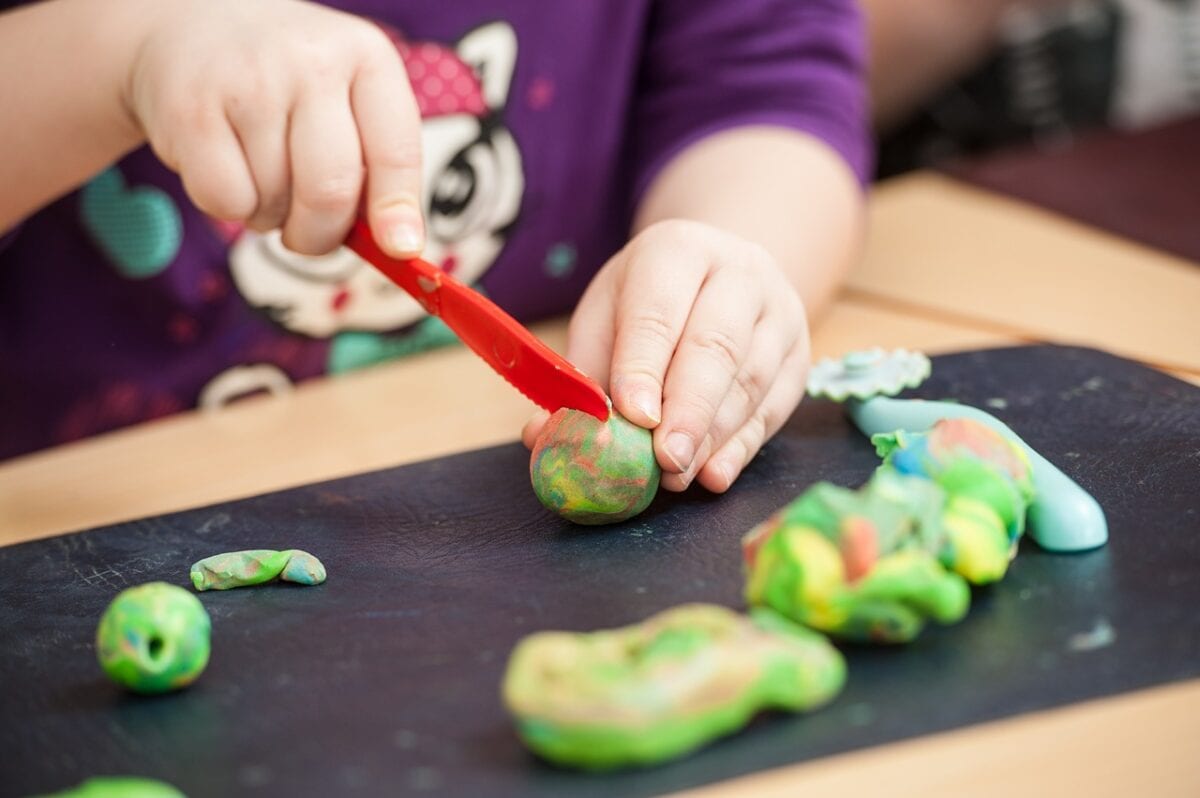 Close-up of a child using a red plastic knife to cut colorful green, orange, and blue playdough on a black mat, with several other playdough creations nearby.